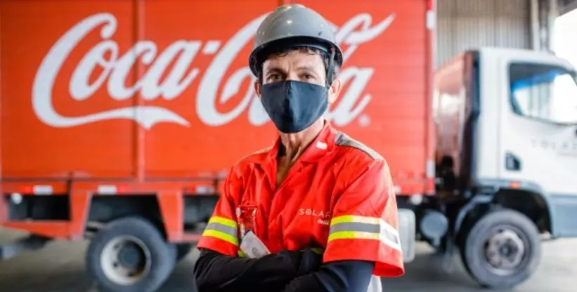 Foto mostra um caminhão da Solar Coca-Cola no fundo e um funcionário da empresa uniformizado, em pé com os braços cruzados e com capacete e máscara