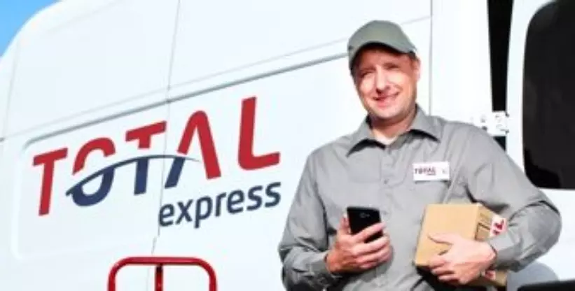Foto mostra um homem segurando uma caixa e um celular ao lado de uma van com o nome da Empresa Total Express
